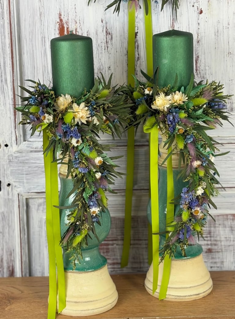 Two green candles in cream vessels with floral garland and lime ribbon against weathered white wood background